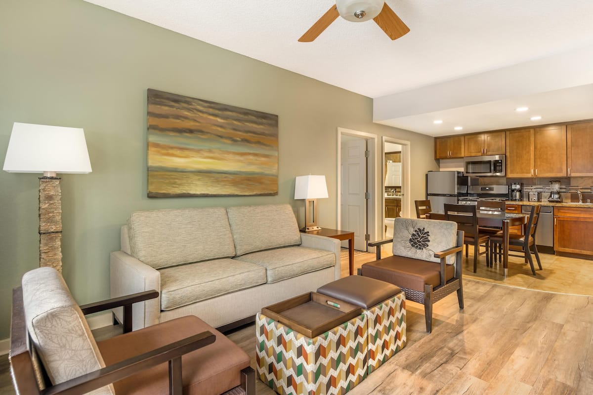 A living room with a sofa, armchairs, a patterned ottoman, a painting, and a ceiling fan.