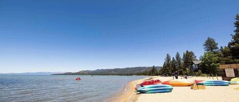 Una playa con kayaks alineados en la arena, cielo despejado y un lago tranquilo al fondo.