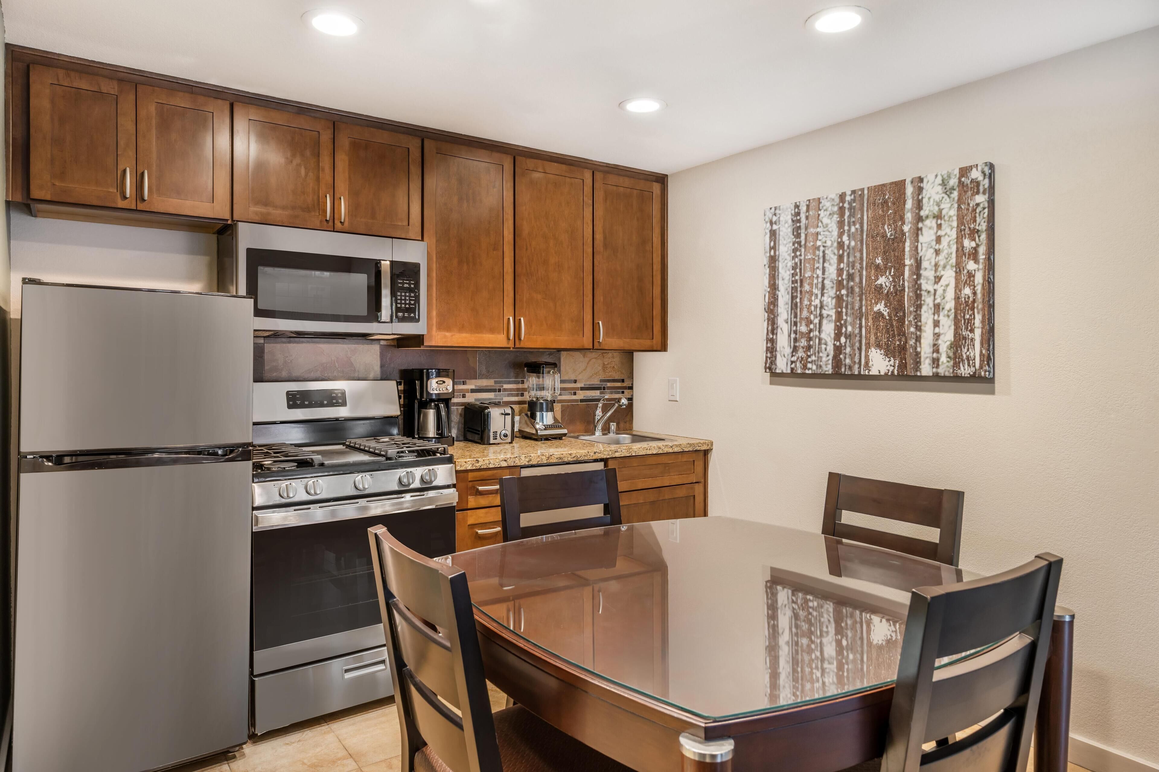 A modern kitchen with stainless steel appliances, wooden cabinets, a glass dining table, and a decorative wall piece.