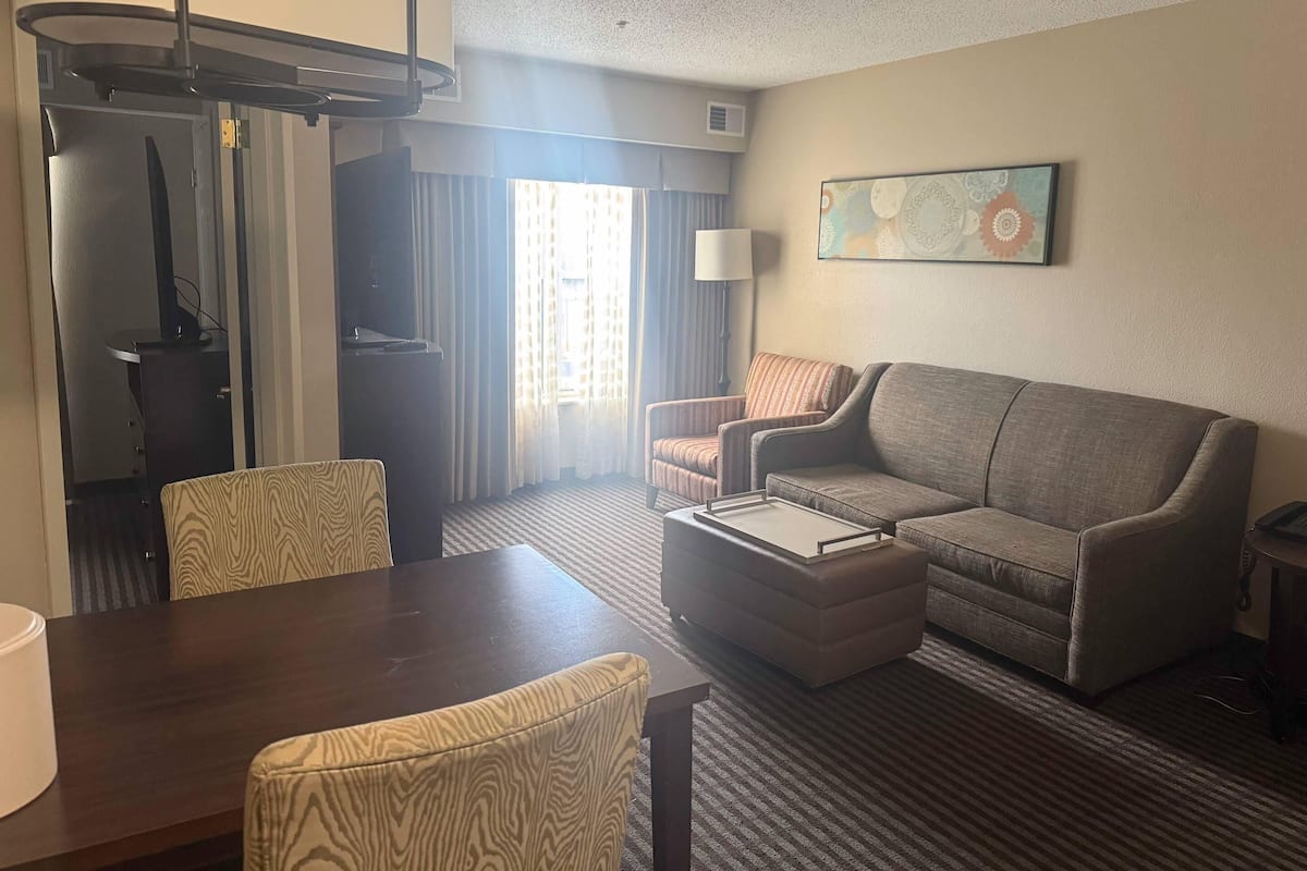 A hotel room with a sofa, armchair, dining table, and a painting on the wall.