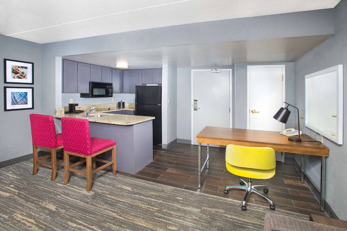 A compact kitchen with a microwave, refrigerator, and granite countertop. There are two chairs with red and yellow upholstery, a wooden desk, and a yellow office chair. The room has a carpeted floor and light gray walls.