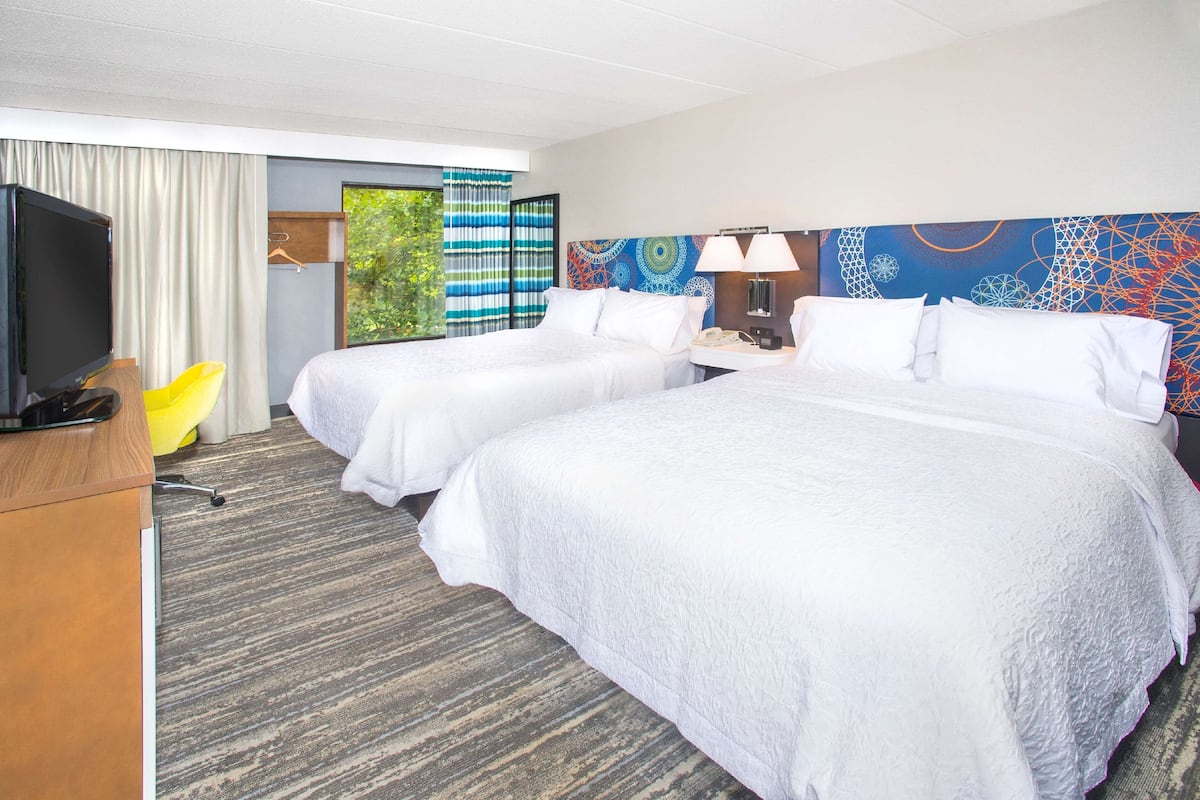 A hotel room with two beds, a TV, a desk, and a window with curtains.