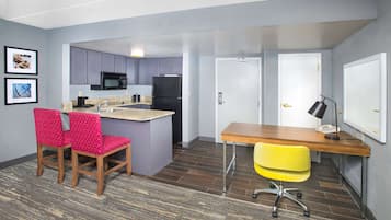 A compact kitchen with a microwave, refrigerator, and granite countertop. There are two chairs, one red and one yellow, and a wooden desk with a lamp.