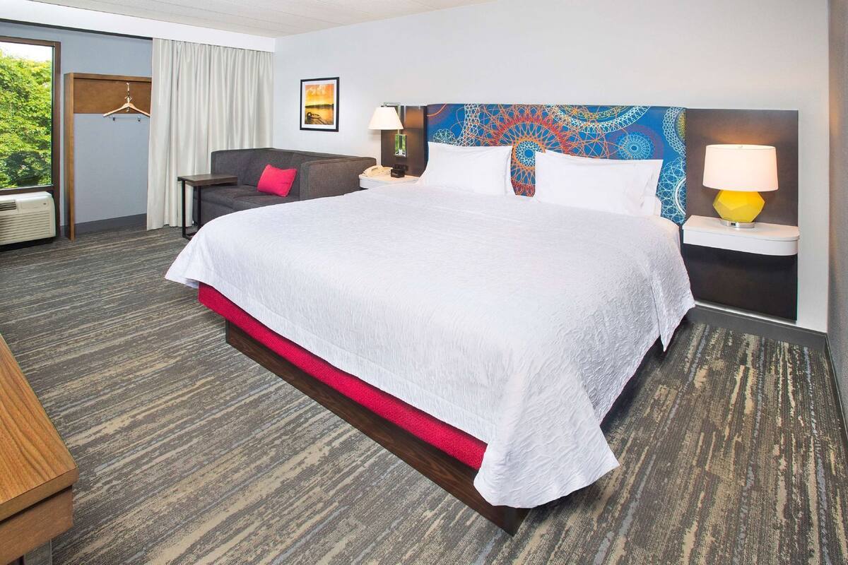 A hotel room with a large bed, a desk, a chair, a lamp, and a view of greenery outside.