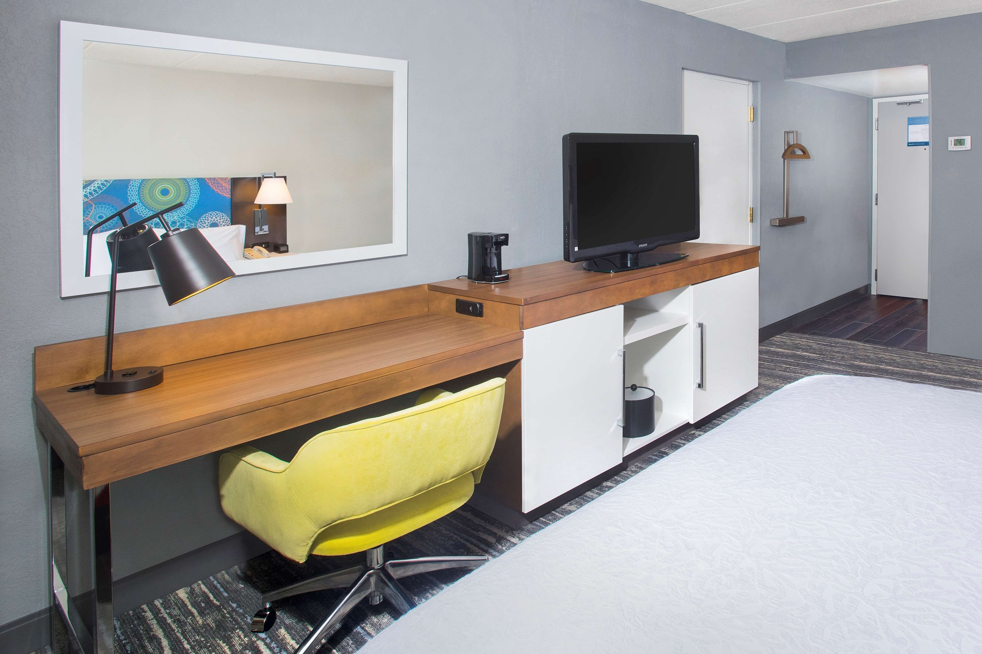 A hotel room with a wooden desk, a television, a yellow chair, a bed, and a large mirror.