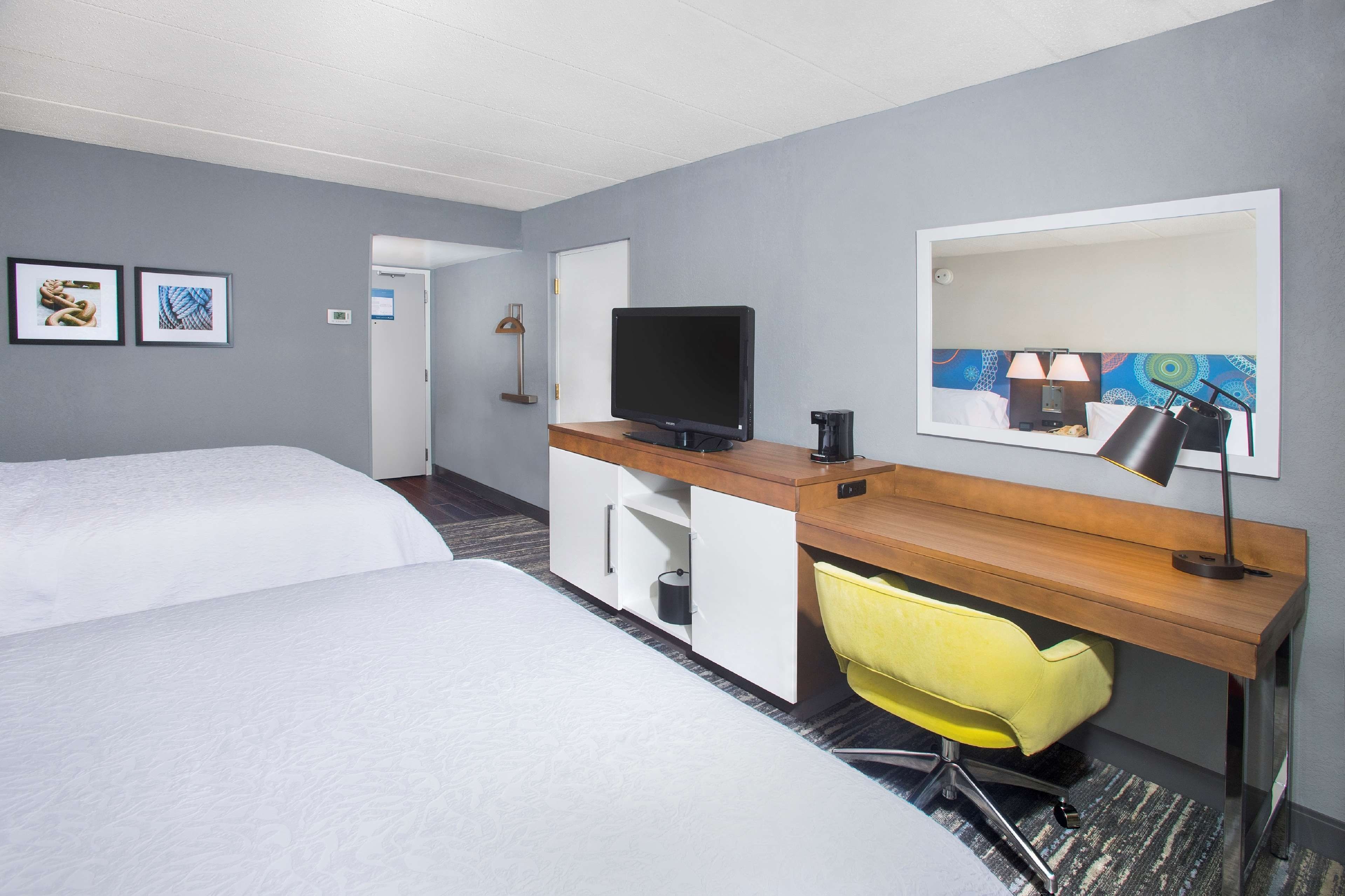 A hotel room with two beds, a desk with a computer, a chair, a desk lamp, and a mirror.