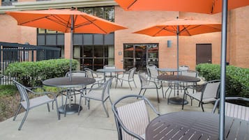Un espace extérieur aménagé en terrasse, avec des tables et des chaises, protégé par de grands parasols orange, et un bâtiment en briques en arrière-plan.