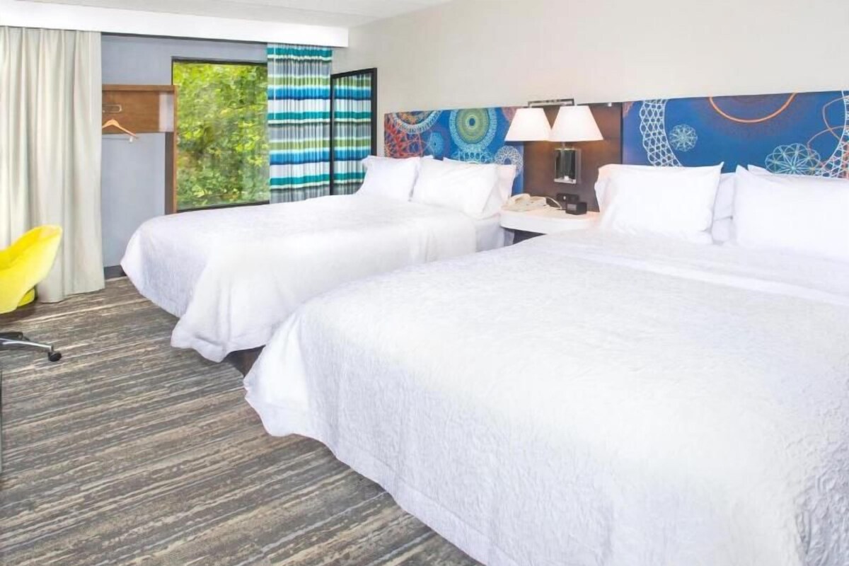 Habitación de hotel con dos camas, un escritorio con lámpara y vista a zonas verdes.