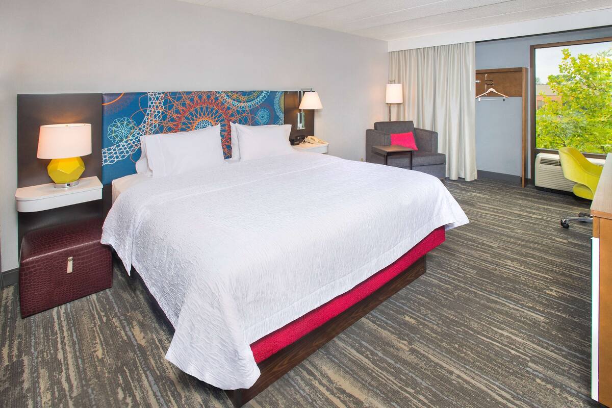 A hotel room with a large bed, a desk with a lamp, a chair, and a view of greenery outside.
