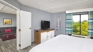 Una habitación de hotel con una cama, un escritorio con televisor, un sofá con almohada roja y una ventana con cortinas.