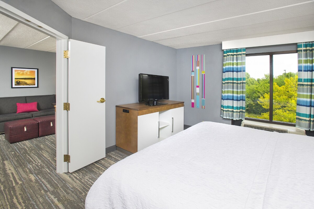 Una habitación de hotel con una cama, un escritorio con televisor, un sofá con almohada roja y una ventana con cortinas.