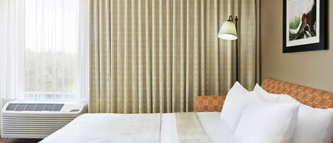 A hotel room with a large bed, an air conditioning unit, a window with sheer curtains, a patterned headboard, a bedside lamp, and a framed picture on the wall.