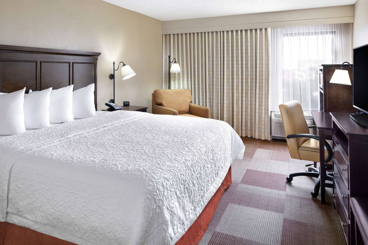 A hotel room with a large bed, a desk with a chair, a television, and a window with curtains.