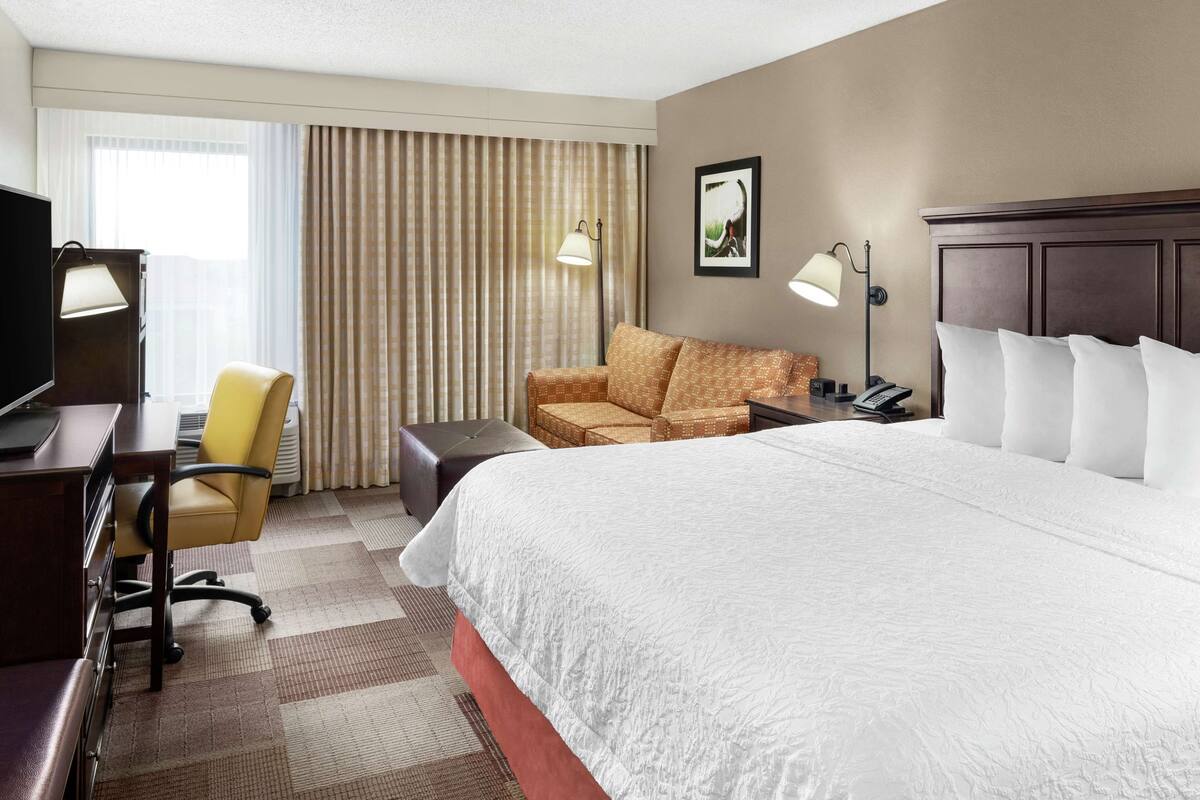 A hotel room with a large bed, a desk with a lamp, a chair, a sofa, and a TV.