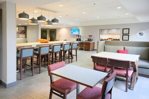 A modern hotel dining area with a long communal table, individual dining tables, and a seating area with a sofa and chairs.