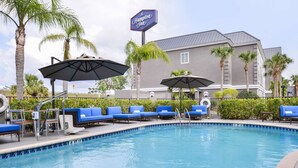 Area piscina con sedie a sdraio blu e ombrelloni, un cartello con la scritta Hampton Inn e un edificio sullo sfondo.