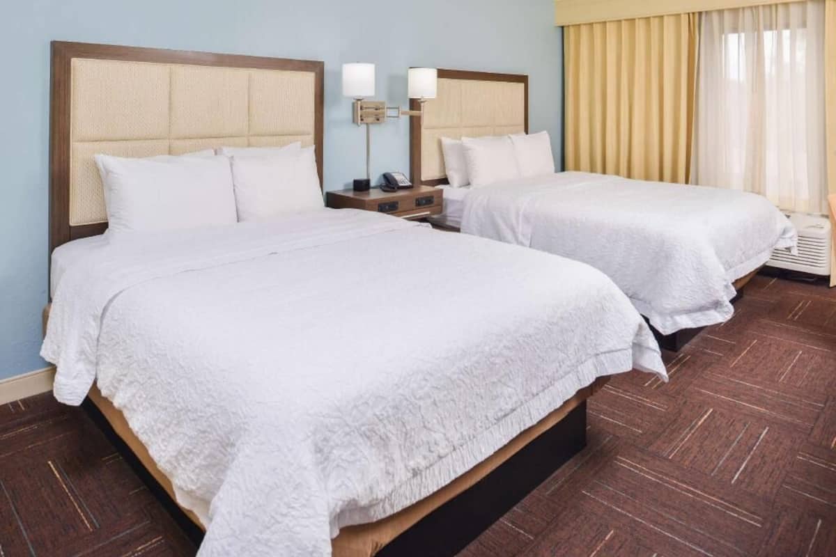 A hotel room with two beds, a nightstand with a lamp, and a window with curtains.