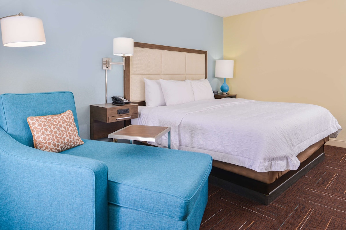 A hotel room with a bed, a blue armchair, a bedside table with a lamp, and a nightstand with a phone.