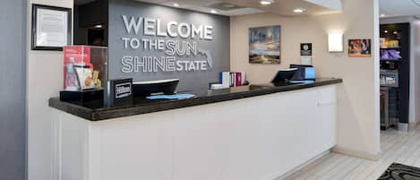 A hotel reception desk with a 'Welcome to the Sun State' sign, a framed certificate, and a display of bottled water and snacks.