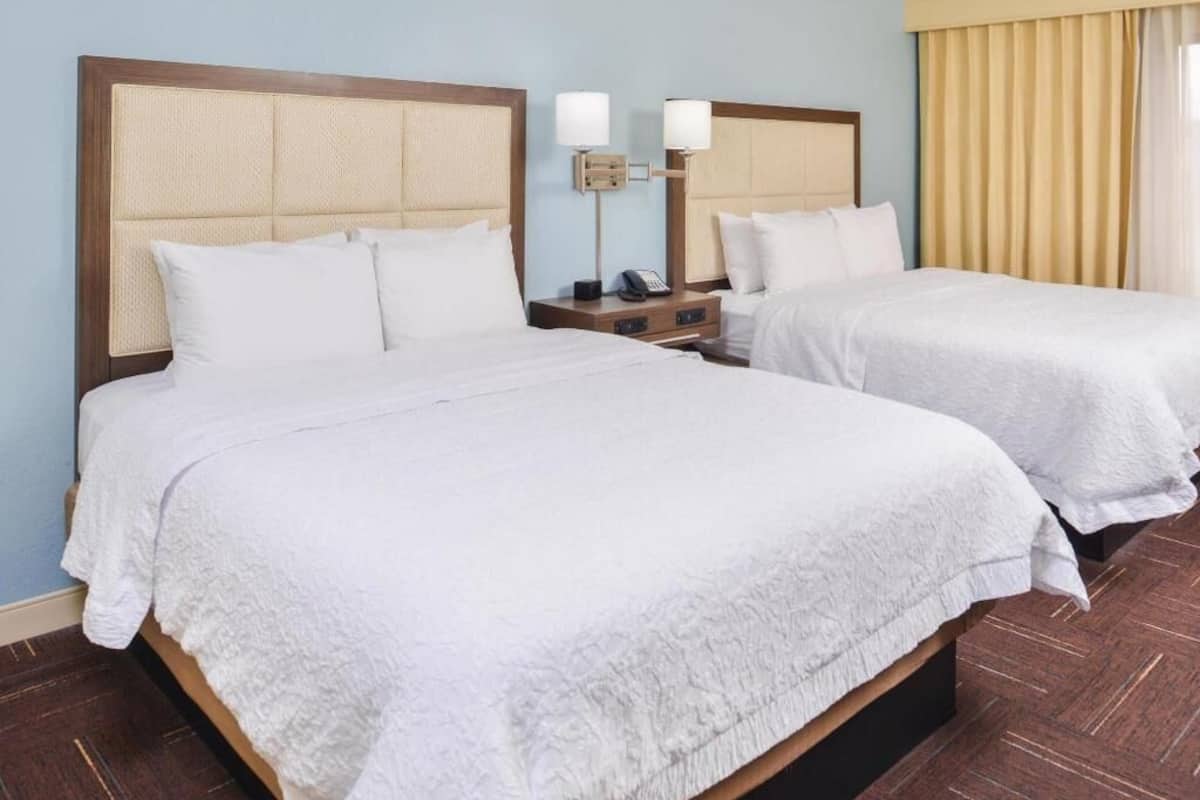 A hotel room with two beds, a nightstand with a lamp, and a window with curtains.