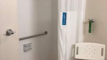 Un espace douche avec un rideau de douche blanc, une barre d’appui, une chaise blanche et une petite étagère.