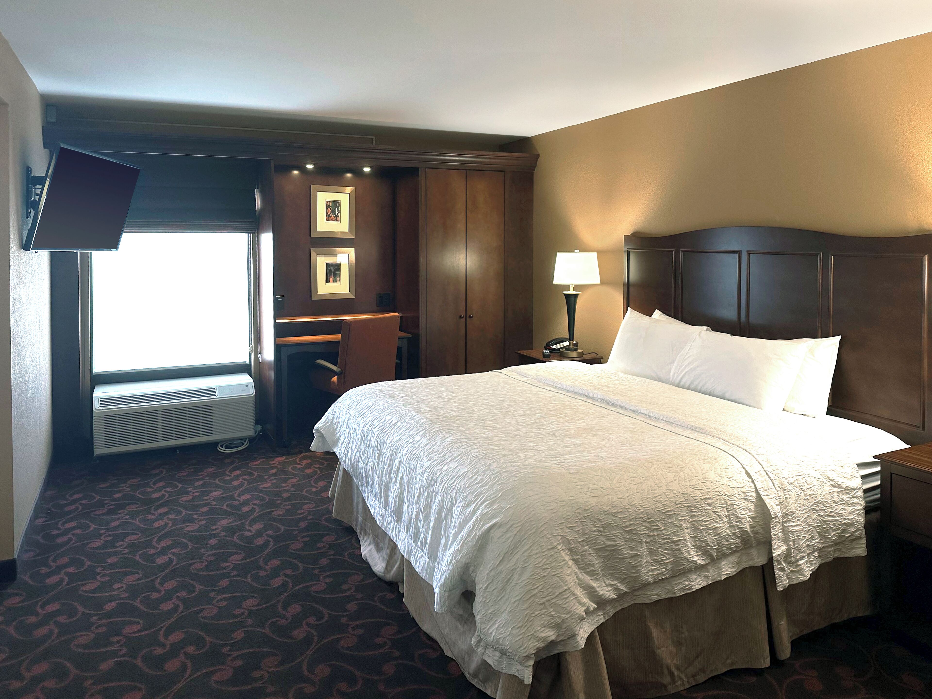 Habitación de hotel con una cama grande, un escritorio, una silla, una lámpara de escritorio, un teléfono y un televisor instalado en la pared.
