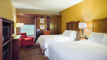 A hotel room with two beds, a desk with a chair, a television, and a window with a view of a green landscape.