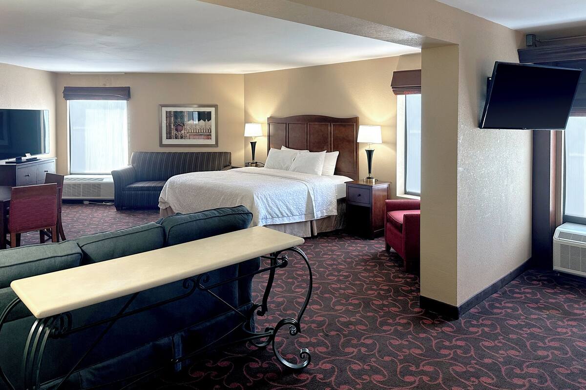 A hotel room with a bed, a desk with a computer, a TV, and a seating area with a bench and chairs.