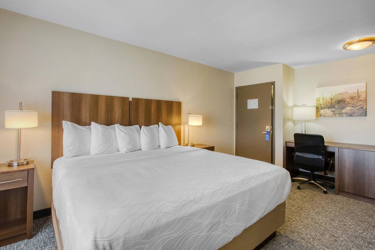 A hotel room with a large bed, two bedside lamps, a desk with a chair, and a door.