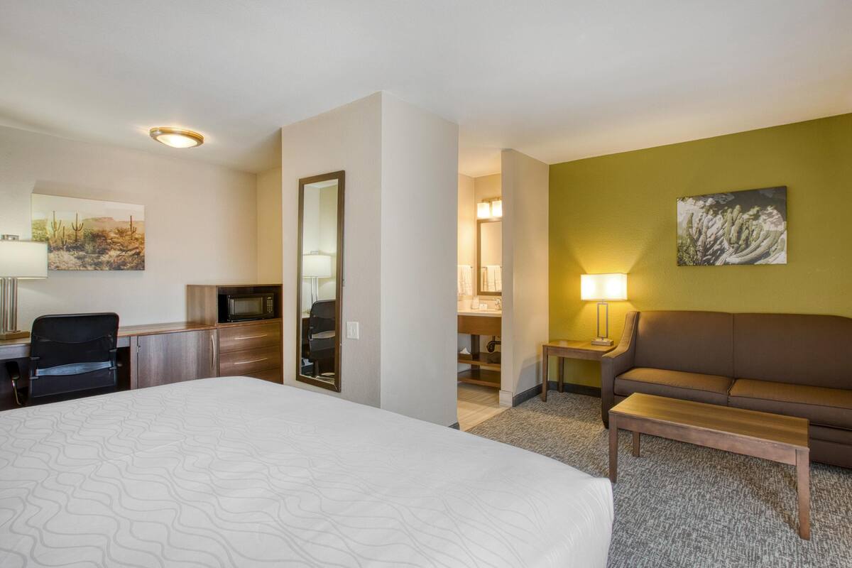 A hotel room with a large bed, a desk with a chair, a sofa, a small table, and a lamp. There is a mirror on the wall and artwork above the sofa.