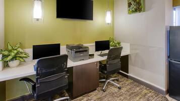 A conference room with two desks, each with a computer monitor, keyboard, and printer. There are two chairs, a flat-screen TV mounted on the wall, and a potted plant on the desk.