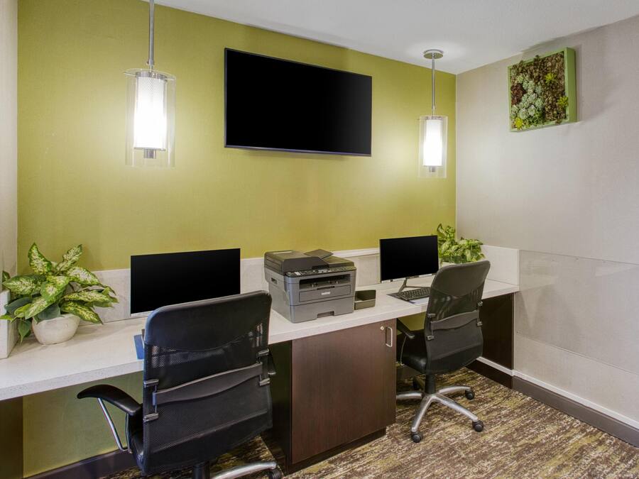 A conference room with two desks, each with a computer monitor, keyboard, and printer. There are two chairs, a flat-screen TV mounted on the wall, and a potted plant on the desk.