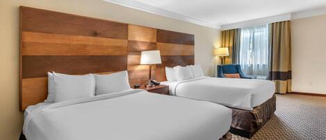 Habitación de hotel con dos camas, cabecera de madera, escritorio con lámpara y vista al exterior a través de una ventana con cortinas.