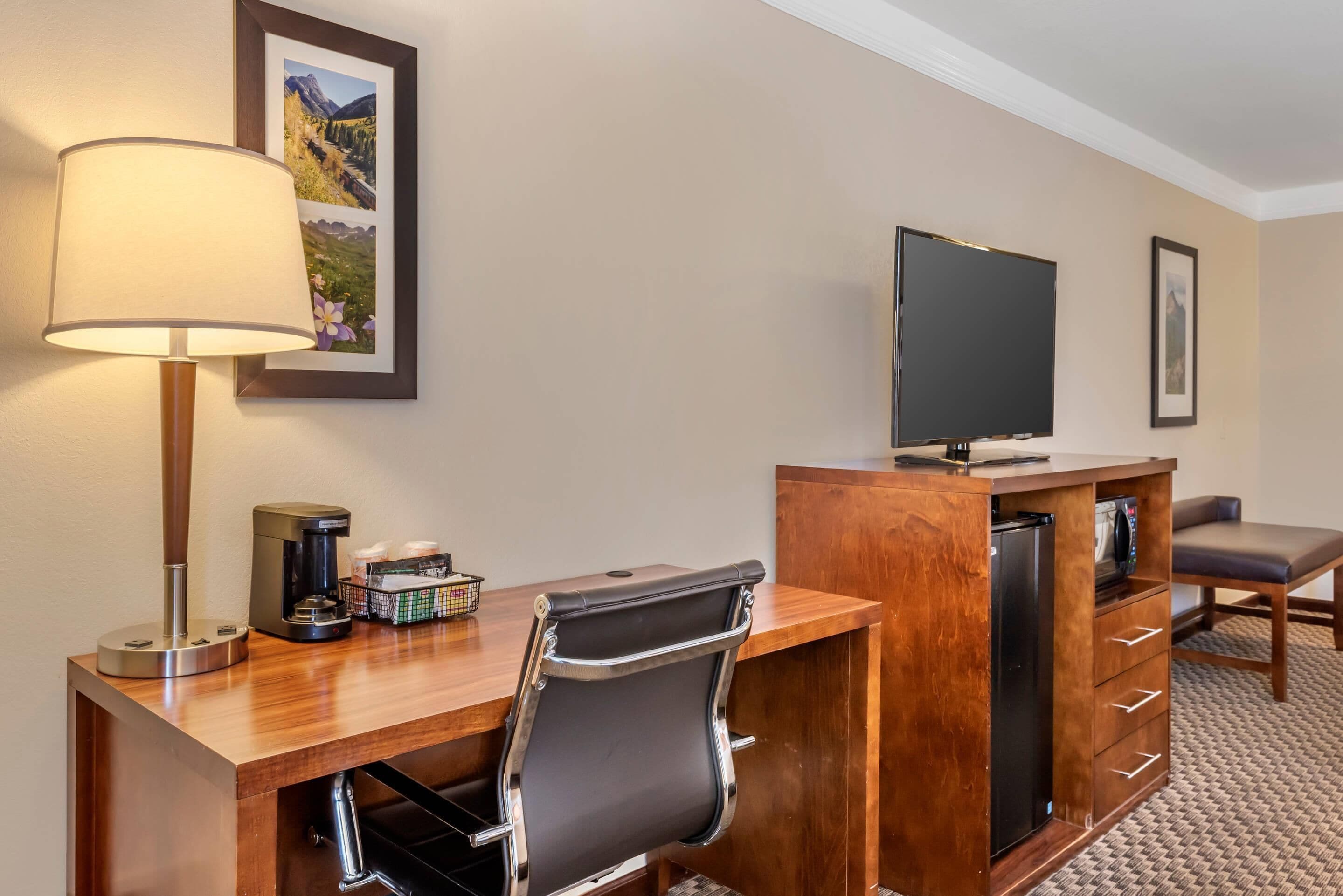 A hotel room with a wooden desk, a black leather chair, a television, a coffee maker, and a refrigerator.