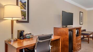 A hotel room with a wooden desk, a black leather chair, a television, a coffee maker, and a refrigerator.