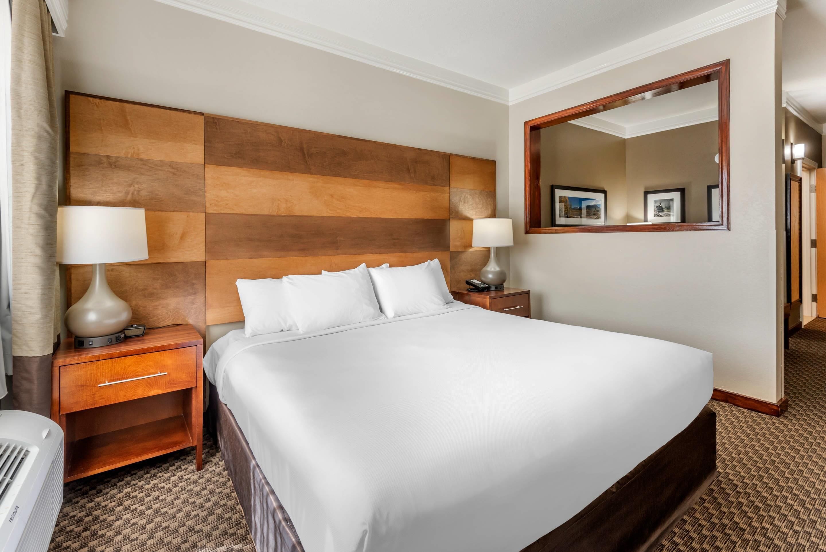 Een hotelkamer met een groot bed, een houten hoofdbord, twee nachtkastjes met lampen, een spiegel en een vloerbedekking.