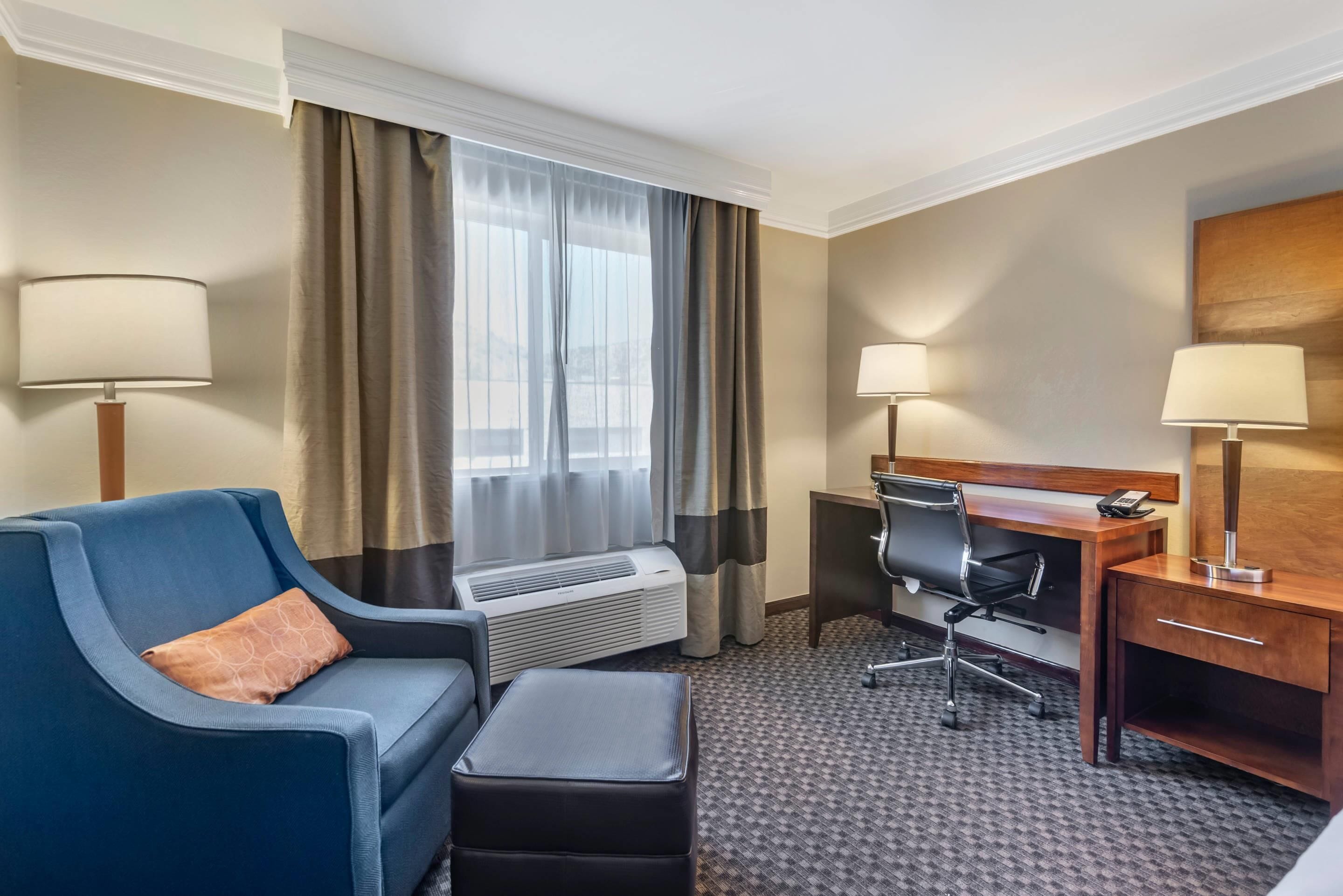 A hotel room with a blue sofa, a desk with a chair, a small table, a lamp, and a large window with curtains.