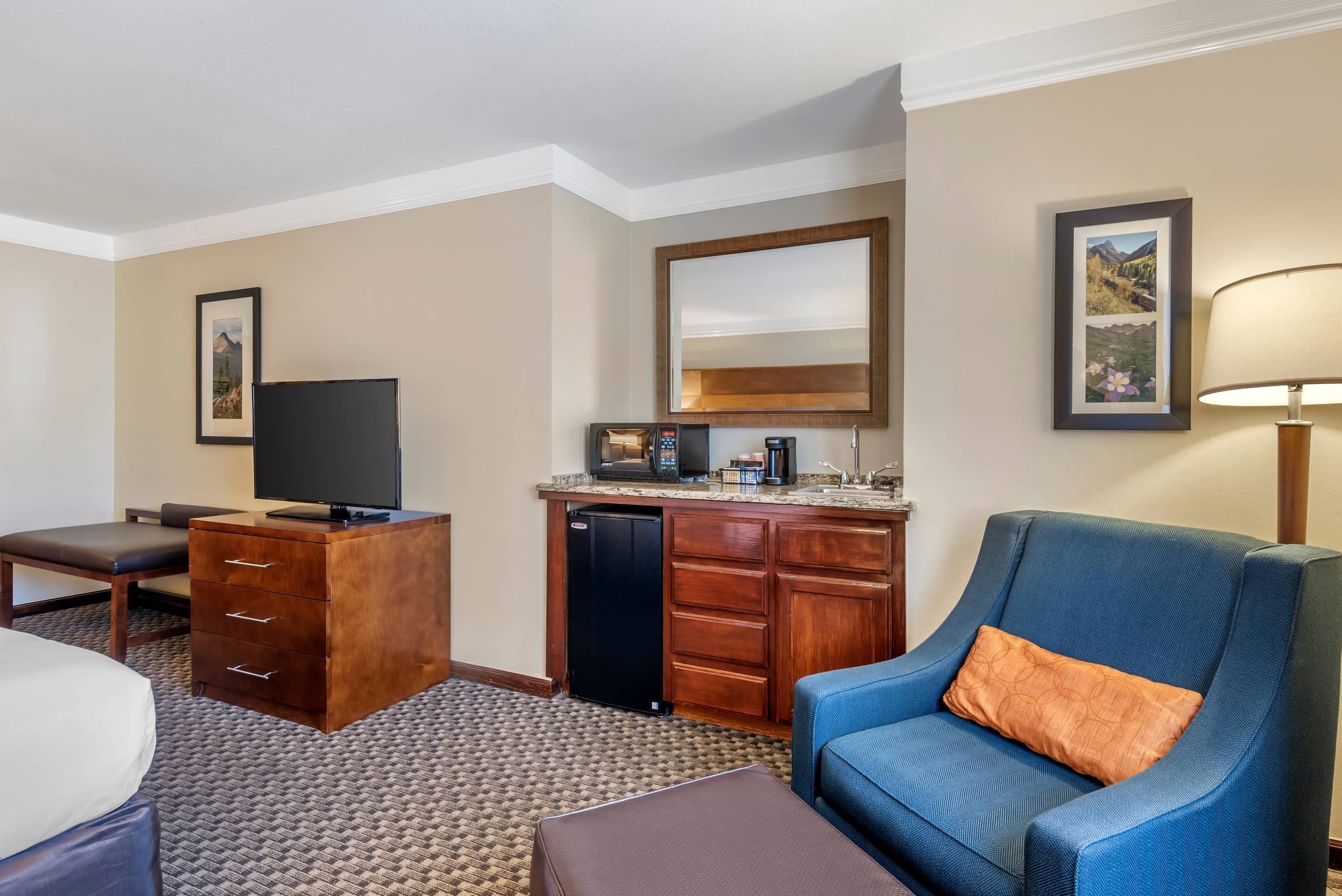 Habitación de hotel con cama, escritorio, silla, televisor, refrigerador, microondas, cafetera y lámpara.
