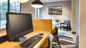 Ein moderner Arbeitsraum mit einem großen Bildschirm, Tastatur und einem ergonomischen Stuhl. Ein Fenster bietet Blick auf Bäume, und an der Wand hängt ein Bild.