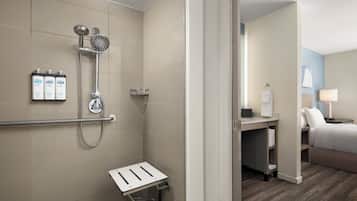 Baño de hotel con ducha, barra de apoyo y un taburete pequeño.