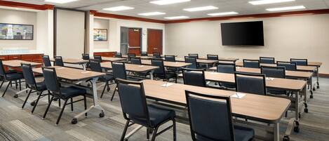 A conference room with multiple tables and chairs, a flat-screen TV mounted on the wall, and a carpeted floor.
