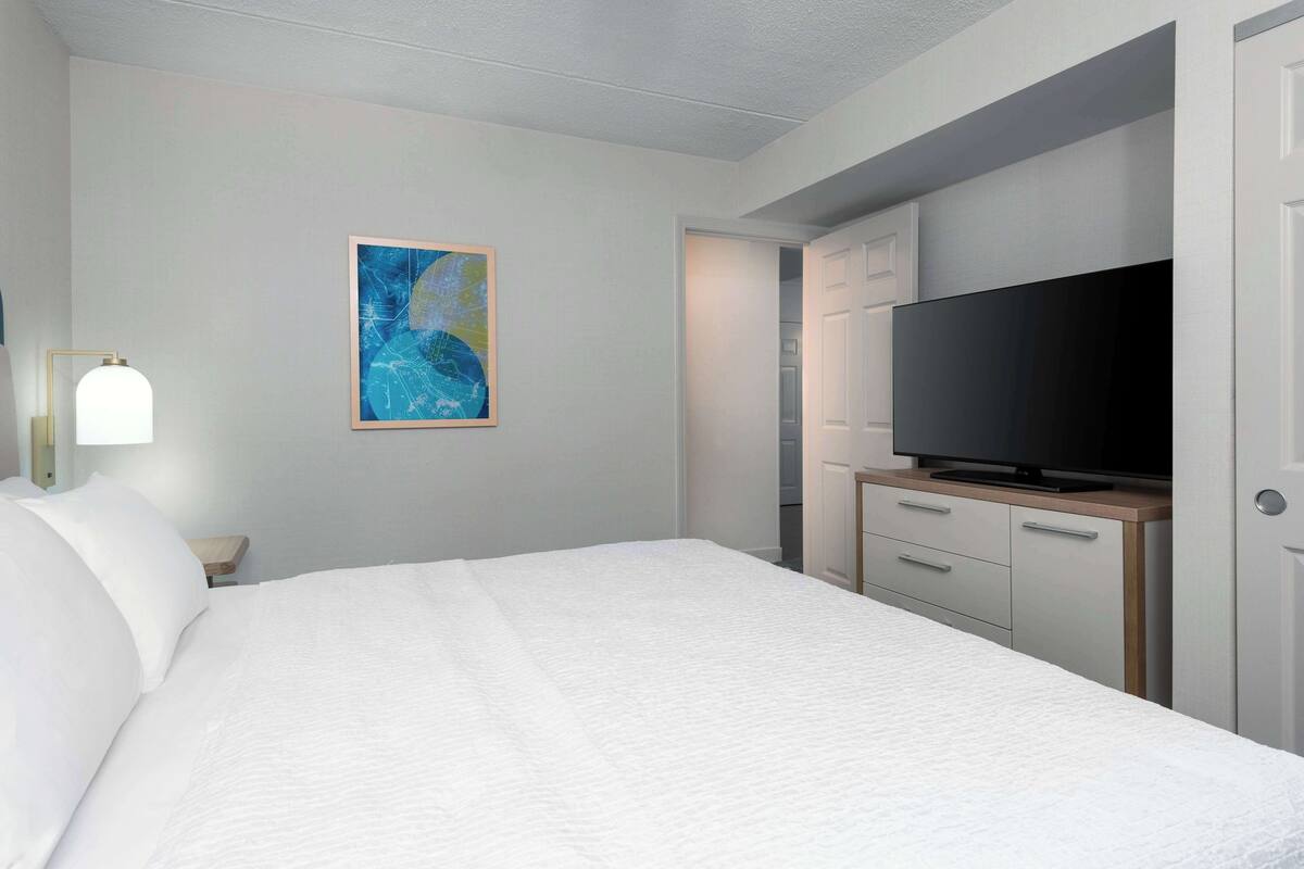 A hotel room with a large bed, a television on a wooden cabinet, and a painting on the wall.
