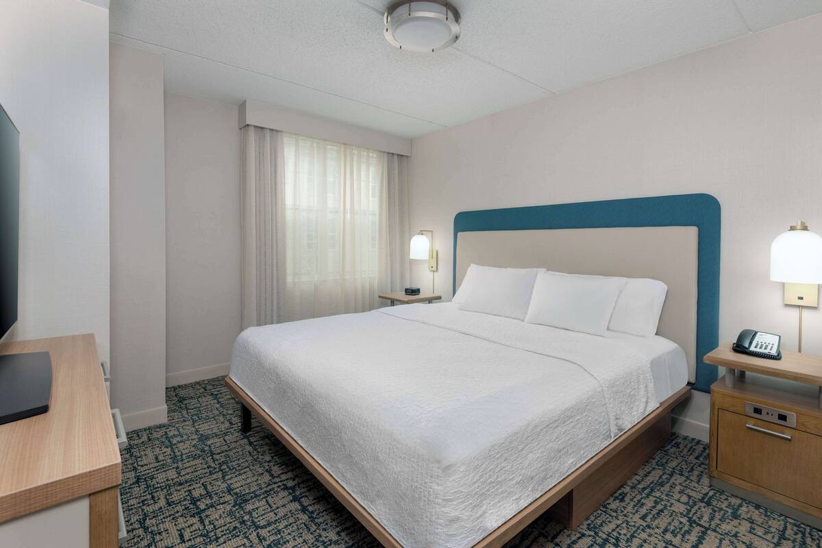 A hotel room with a bed, bedside table, lamp, and a desk with a chair.