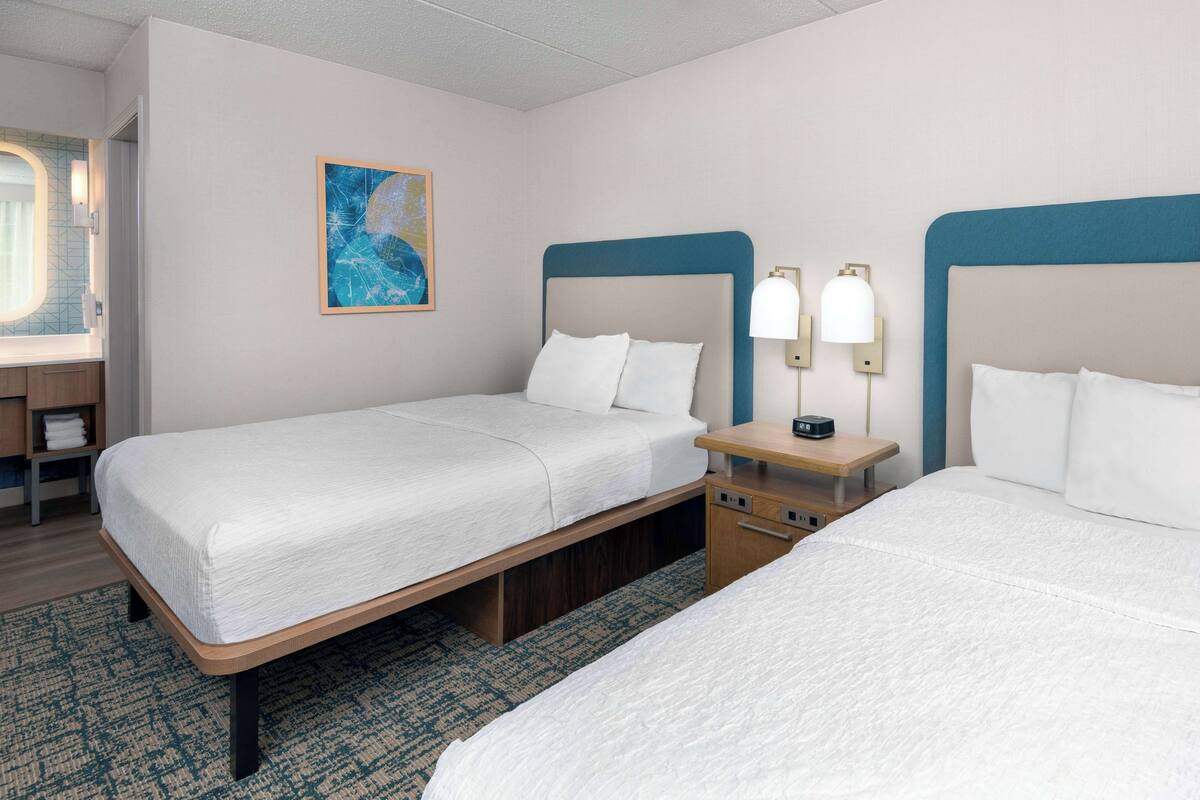 A hotel room with two beds, a nightstand, a lamp, and a painting on the wall.