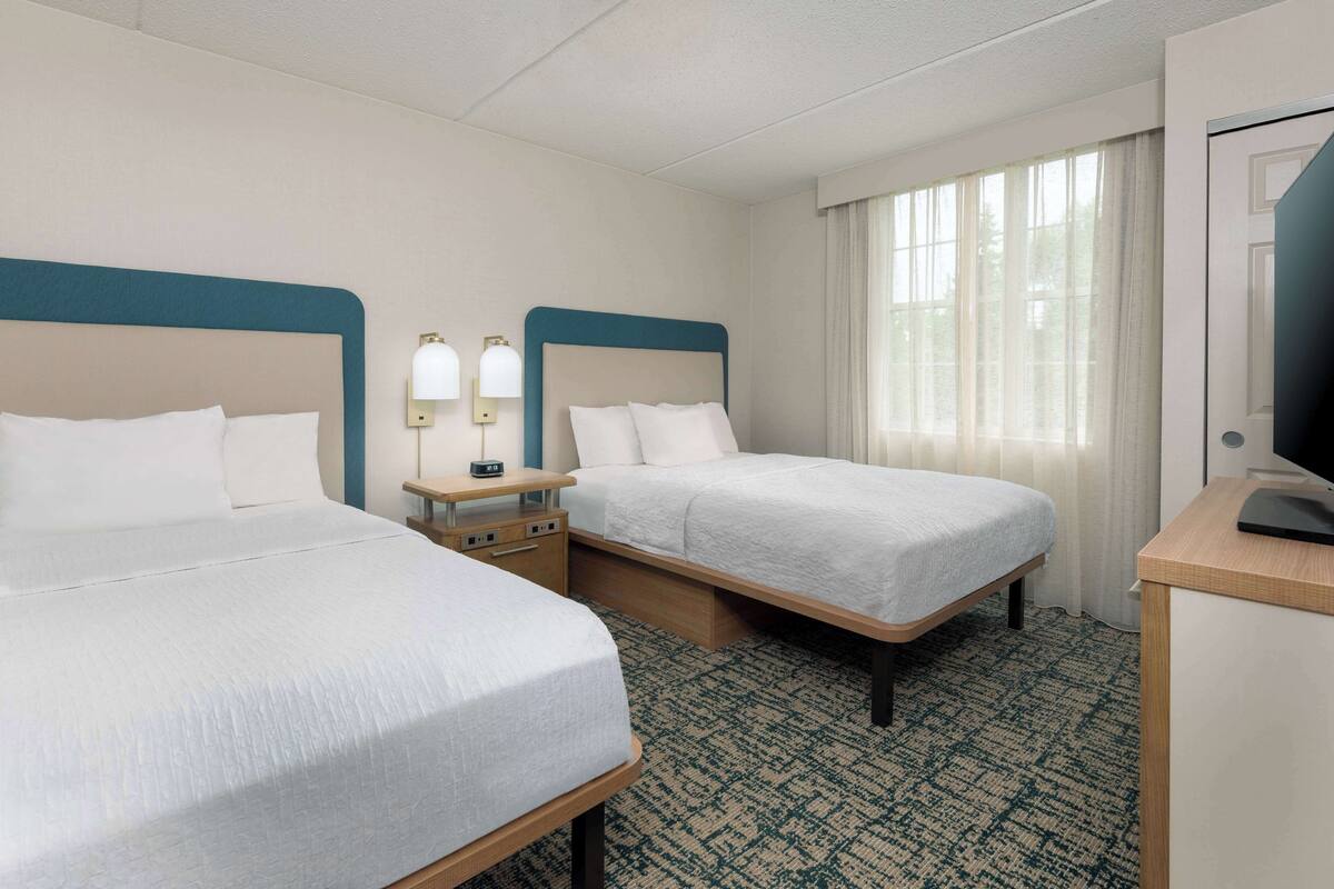 A hotel room with two single beds, a bedside table with a lamp, a desk with a computer, and a window with curtains.