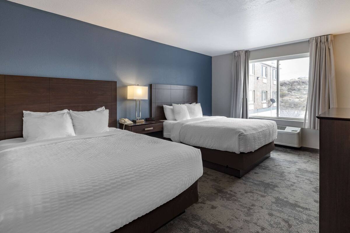A hotel room with two beds, a nightstand, a lamp, and a window with curtains.