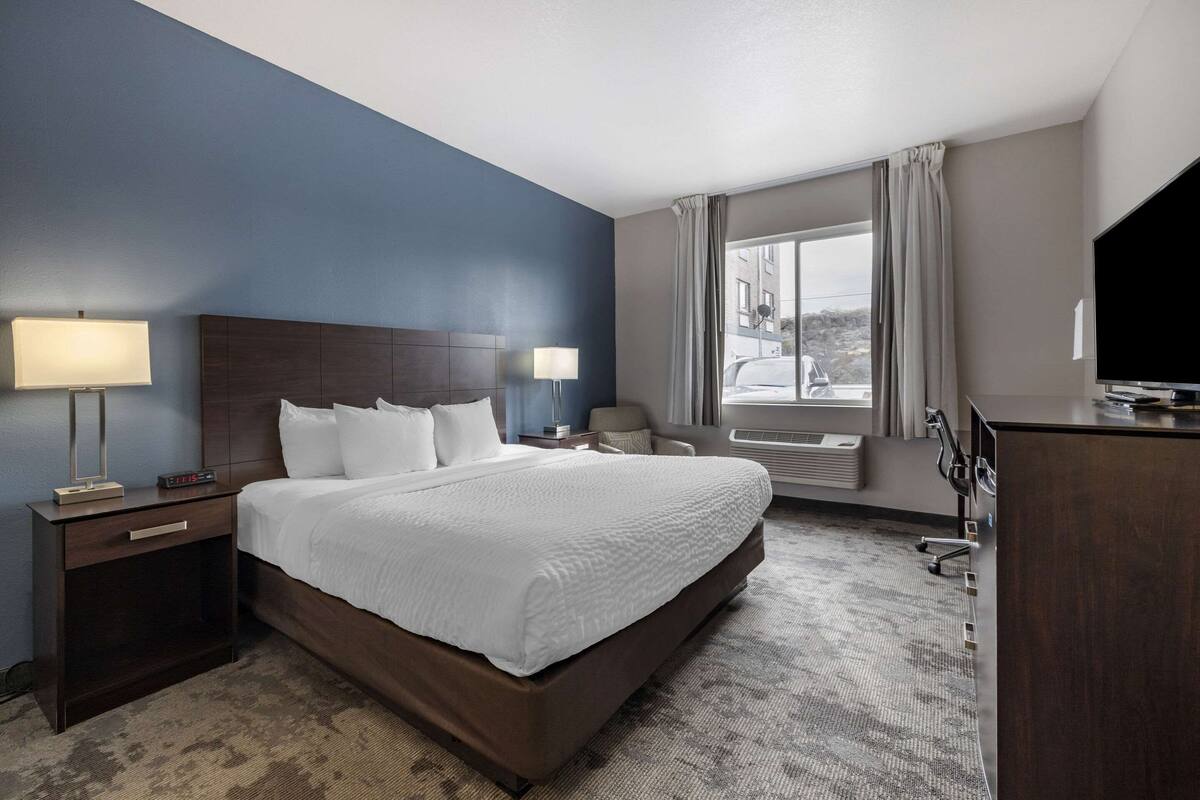 A hotel room with a large bed, two bedside tables with lamps, a desk with a flat-screen TV, and a window with curtains.
