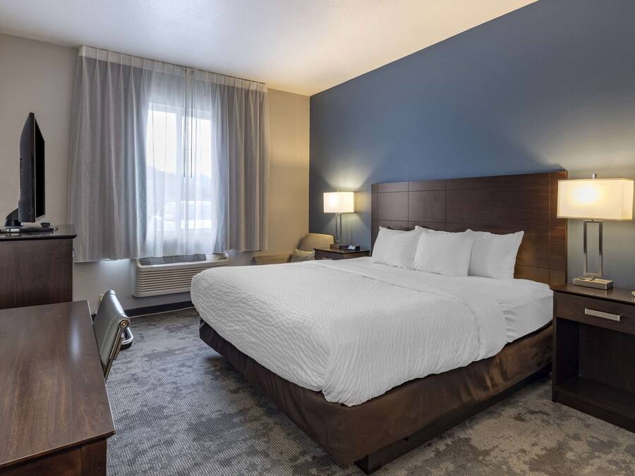 A hotel room with a large bed, two bedside tables with lamps, a desk, a TV, and a window with curtains.