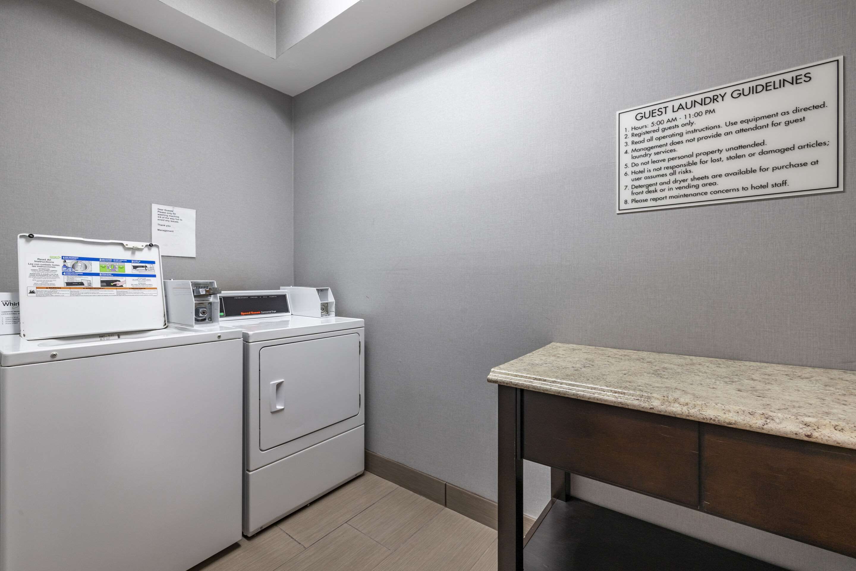 A laundry area with a sign detailing guest laundry guidelines, a washing machine, a dryer, and a small table with a countertop.