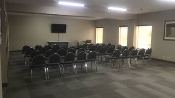 Uma sala de conferências com fileiras de cadeiras, uma TV de tela plana e janelas amplas.